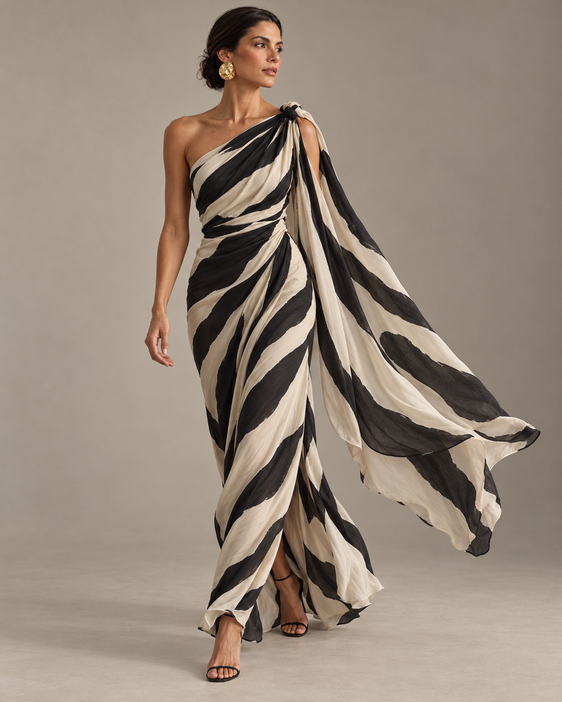 Modelo caminhando com vestido longo de listras pretas e off-white, de um ombro só, com nó no ombro e uma faixa de tecido solta ao vento